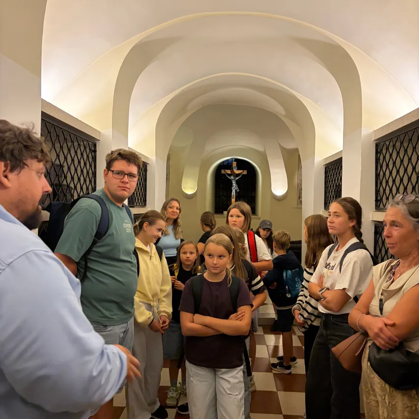 Eine Gruppe von Menschen, darunter Kinder, steht in einem Flur mit gewölbten Decken, möglicherweise in einer Kirche. Ein Kind schaut zur Kamera.