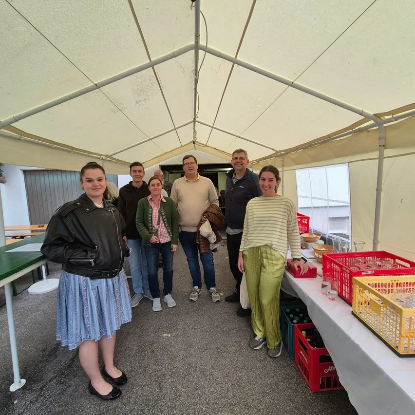 Eine Gruppe von Menschen, sowohl Männer als auch Frauen, stehen in einem Zelt mit einem weißen Dach. Sie lächeln und scheinen für ein Foto zu posieren. Der Tisch hat eine weiße Tischdecke und einige Gegenstände darauf. Es gibt auch einige rote Kisten mit Gläsern und anderen Gegenständen auf dem Tisch.