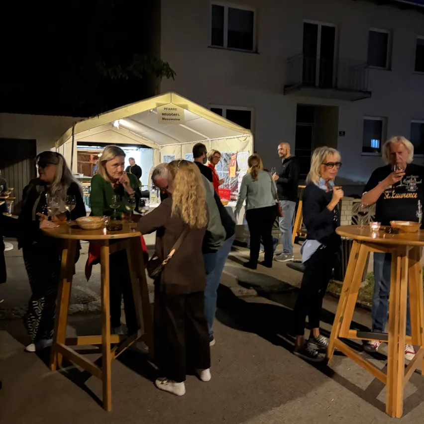 Eine Gruppe von Menschen versammelt sich bei Nacht um Holztische draußen, einige halten Weingläser. Im Hintergrund steht ein Zelt mit einem Schild.
