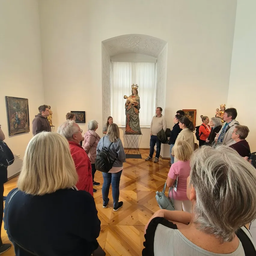 Eine Gruppe von Menschen versammelt sich in einem Museumsraum und bewundert eine Statue einer gekrönten Frau, die ein Kind hält. Der Raum hat Holzböden und weiße Wände, die mit verschiedenen Gemälden geschmückt sind.