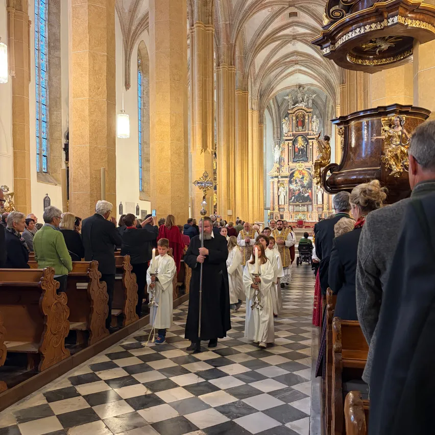 Eine religiöse Prozession in einer Kathedrale, angeführt von einem Priester, der Kinder mit Kerzen führt. Das Kirchenschiff ist mit Besuchern gefüllt und der Altar ist mit kunstvollen Dekorationen geschmückt.