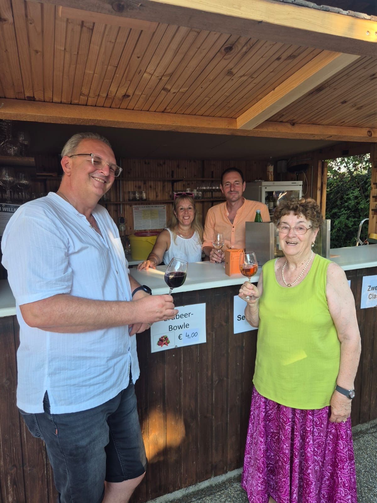 Vier Personen stehen lächelnd an einer Bartheke. Ein Mann links hält ein Glas Wein, ein anderer rechts hält auch eins. Zwei Frauen stehen hinter der Theke.