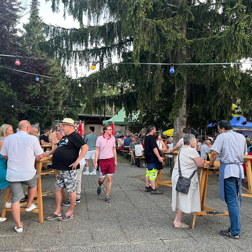 Im Freien eingerichteter Bereich mit Menschen in Hüten, Shirts, Shorts und Sandalen. Es gibt Tische und Stühle. Einige Leute gehen. Der Bereich ist von Bäumen umgeben und hat hängende Lichter.