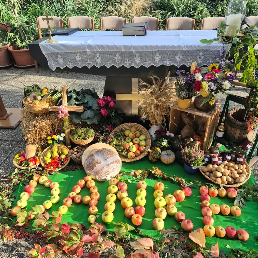 Ein Tisch mit einem weißen Tuch ist im Freien aufgestellt, umgeben von verschiedenen Früchten, Gemüse und Blumen. Ein Holzkreuz und eine Kerze sind auf dem Tisch platziert.