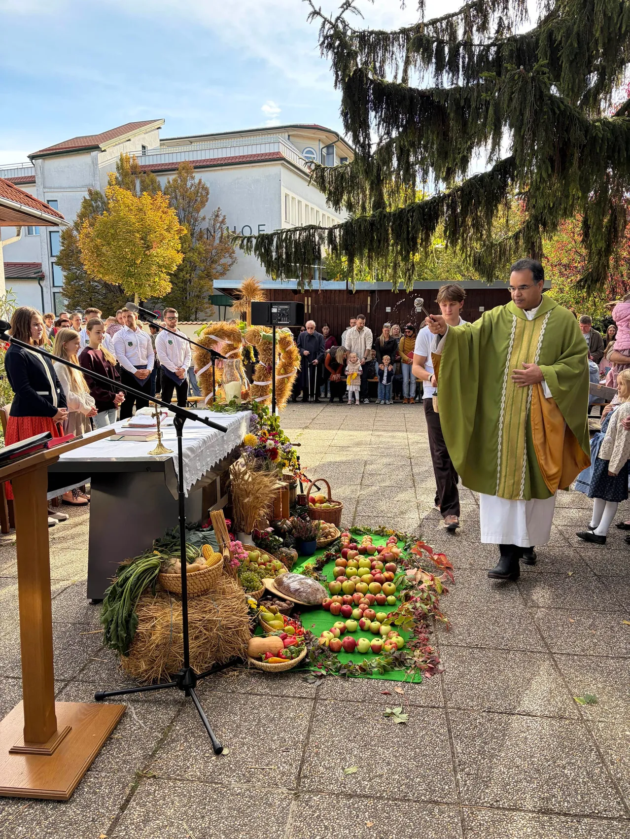 Ein Priester steht vor einem Tisch mit einem Kranz und einem Korb mit Früchten, umgeben von einer Menschenmenge.