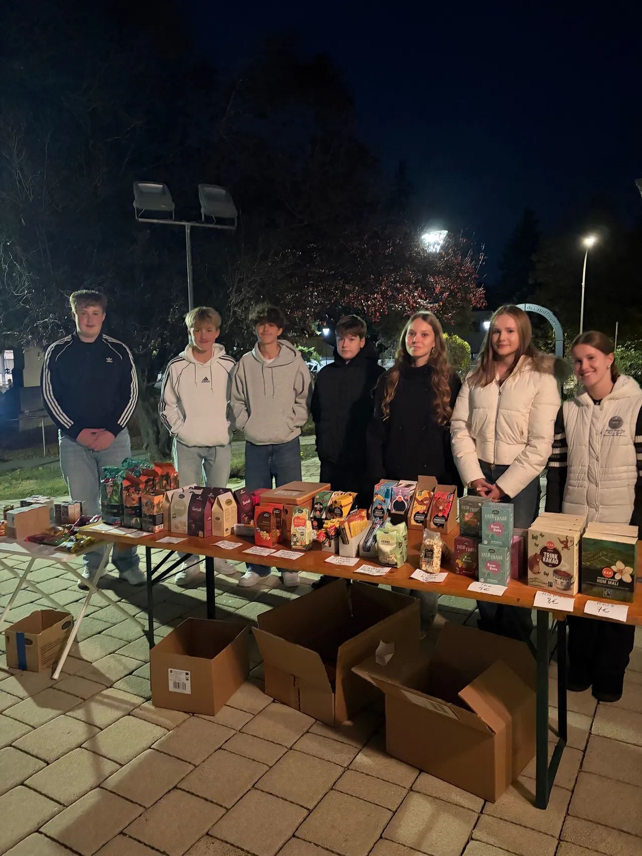 Eine Gruppe junger Leute steht hinter einem Tisch mit verschiedenen Produkten, wahrscheinlich für einen Verkauf, bei Nacht.