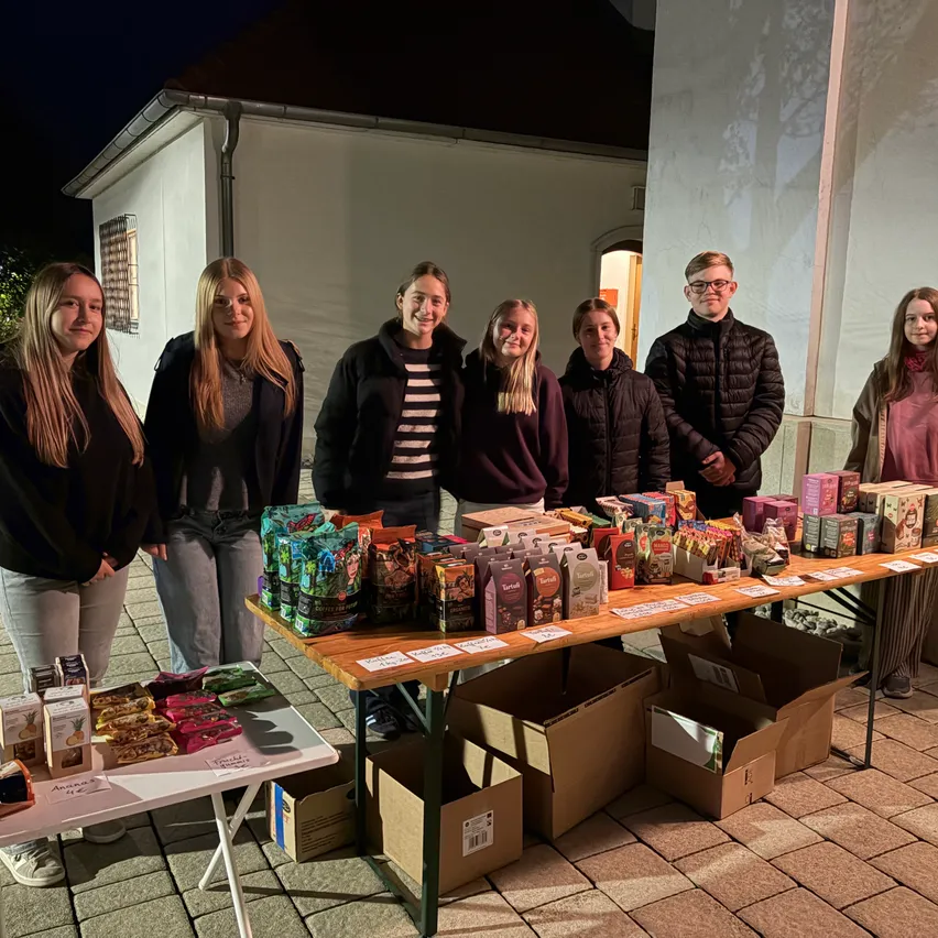 Eine Gruppe junger Menschen steht hinter einem Tisch voller verschiedener Snacks und Kisten, möglicherweise für eine Gemeinschaftsveranstaltung.