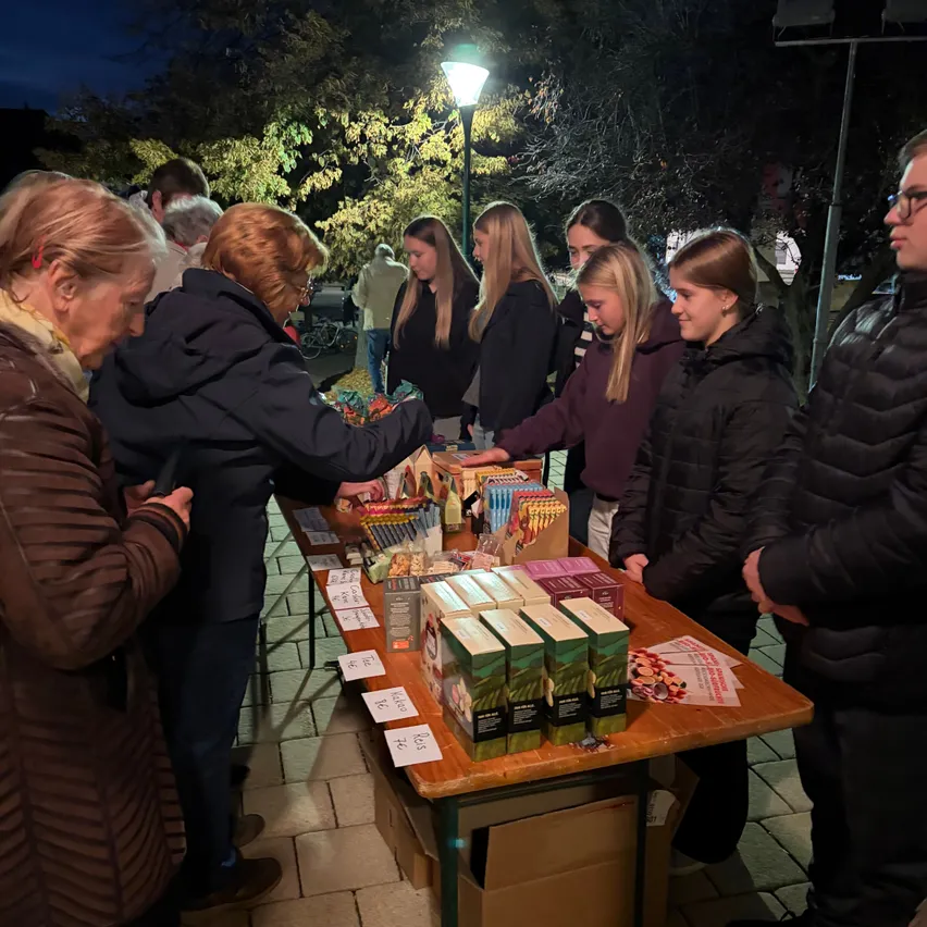 Eine Gruppe von Menschen versammelt sich um einen Tisch mit verschiedenen ausgestellten Gegenständen, wahrscheinlich auf einem Markt oder bei einer Veranstaltung.