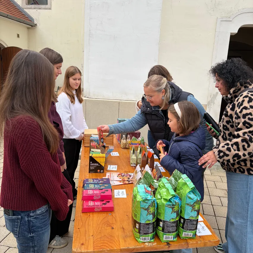 Eine Gruppe von Menschen, einschließlich Erwachsener und Kinder, ist um einen Tisch versammelt, auf dem verschiedene Gegenstände stehen. Zu den Gegenständen gehören Schachteln mit Pralinen, Kaffeesäckchen und andere Gegenstände. Die Menschen betrachten die Gegenstände und scheinen daran interessiert zu sein.