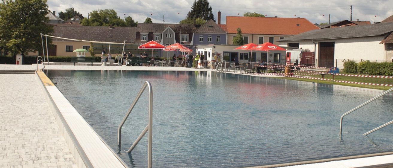 Ein leerer Swimmingpool mit roten Schirmen an der Seite, umgeben von Häusern mit Schornsteinen und einem klaren Himmel mit Wolken.