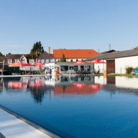 Ein Schwimmbecken spiegelt die Umgebung wider. Gebäude mit roten Dächern und mehrere rote Sonnenschirme sind im Hintergrund zu sehen. Bäume und Büsche sind in der Ferne.