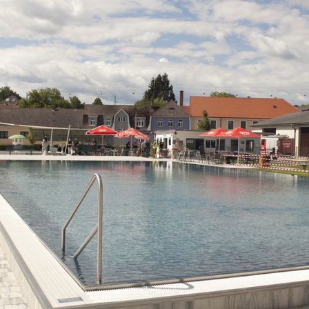 Ein Freibad mit klarem Wasser, einem silbernen Handlauf und mehreren roten Sonnenschirmen. Im Hintergrund befinden sich mehrere Gebäude, und einige Personen sitzen.
