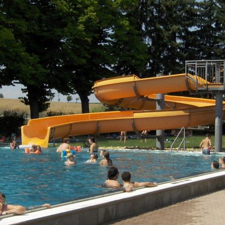 Ein Freibad mit mehreren schwimmenden Personen. Ein orangefarbener Wasserrutsche ist über dem Pool positioniert. Bäume und Gras umgeben das Gebiet.