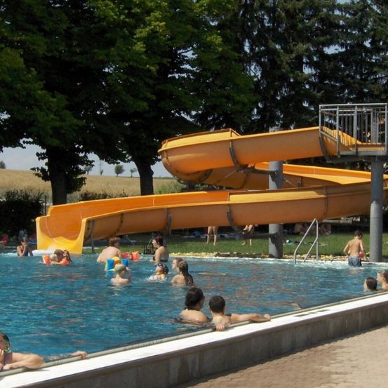 Ein Freibad mit mehreren schwimmenden Personen. Ein orangefarbener Wasserrutsche ist über dem Pool positioniert. Bäume und Gras umgeben das Gebiet.