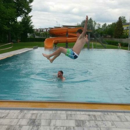 Ein Mann springt in ein Schwimmbecken, während ein anderer Mann darunter schwimmt. Im Hintergrund befindet sich eine orangefarbene Wasserrutsche.