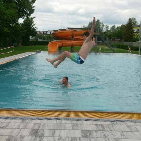 Ein Mann springt in ein Schwimmbecken, während ein anderer Mann darunter schwimmt. Im Hintergrund befindet sich eine orangefarbene Wasserrutsche.