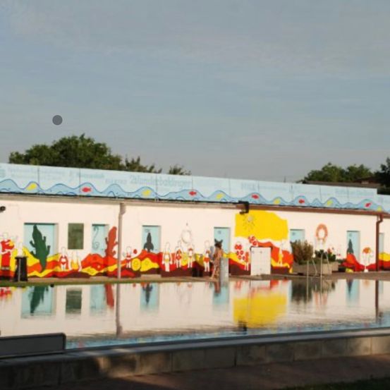 Ein Gebäude mit farbenfrohen Wandgemälden in der Nähe eines Pools mit Reflexionen des Gebäudes, der Bäume und des Himmels. Eine Person steht in der Nähe des Pools.