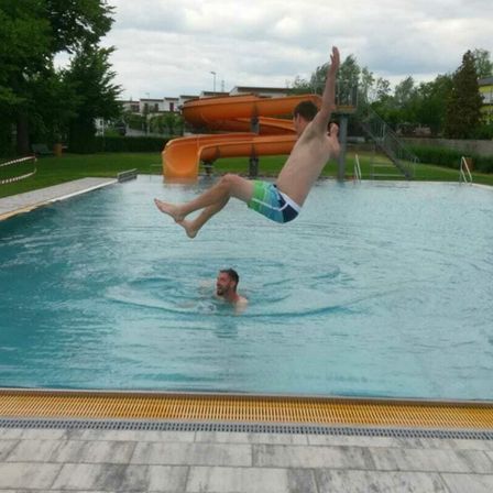 Ein Mann springt in ein Schwimmbecken, während ein anderer Mann darunter schwimmt. In der Nähe befindet sich eine orangefarbene Wasserrutsche.