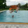 A man jumps into a swimming pool while another man swims below. Nearby, there is an orange water slide.