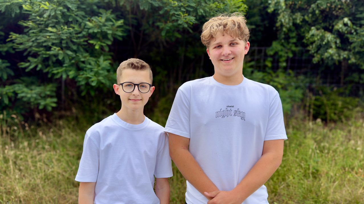 Two young boys stand side by side in a grassy area, both wearing white t-shirts. The boy on the left has glasses and a serious expression, while the boy on the right has a smile.