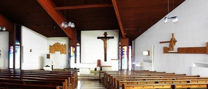 Ein leerer Kircheninnenraum mit Holzbänken, einem Holzkreuz an der Wand und dekorativen Elementen. Die Decke hat Holzbalken und hängende Lampen.