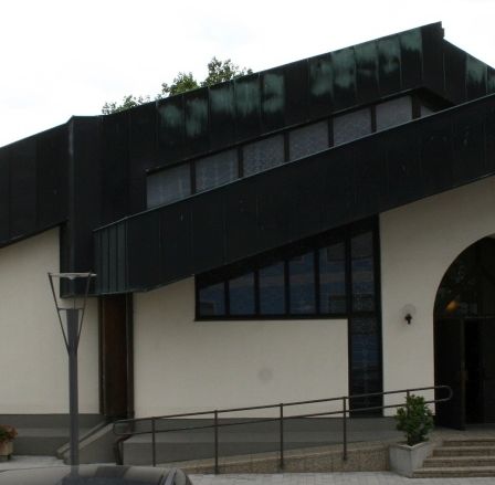 Ein modernes Gebäude mit schwarzem Dach und weißen Wänden. Ein Flagge und eine Pflanze stehen in der Nähe des Eingangs. Ein Auto ist vor dem Gebäude geparkt.