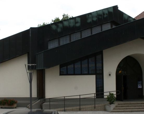 Ein modernes Gebäude mit schwarzem Dach und weißen Wänden. Ein Flagge und eine Pflanze stehen in der Nähe des Eingangs. Ein Auto ist vor dem Gebäude geparkt.