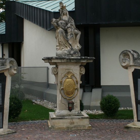 Eine Statue einer Frau mit Krone steht vor einem Gebäude mit grünem Dach. Zwei schwarze Tafeln mit Inschriften flankieren die Statue.