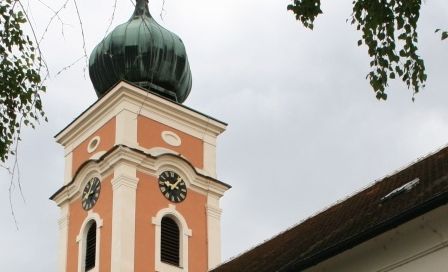 Eine alte Kirche mit orangefarbener Außenfassade und einem grün bedachten Turm. Das Gebäude hat bogenförmige Fenster und eine Statue an der Wand.