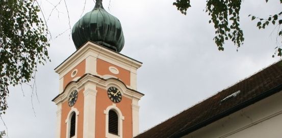 Eine alte Kirche mit orangefarbener Außenfassade und einem grün bedachten Turm. Das Gebäude hat bogenförmige Fenster und eine Statue an der Wand.