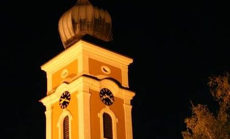 Ein Kirchturm mit einer Kuppel und zwei Uhren ist bei Nacht beleuchtet. Er ist von Bäumen umgeben und hat eine helle Beleuchtung an der Seite.