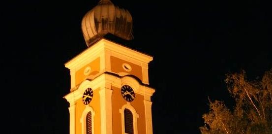 Ein Kirchturm mit einer Kuppel und zwei Uhren ist bei Nacht beleuchtet. Er ist von Bäumen umgeben und hat eine helle Beleuchtung an der Seite.