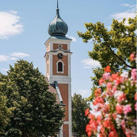 Eine Kirche mit Glockenturm steht unter klarem Himmel, umgeben von Bäumen und Blumen, mit einem geparkten Auto in der Nähe.