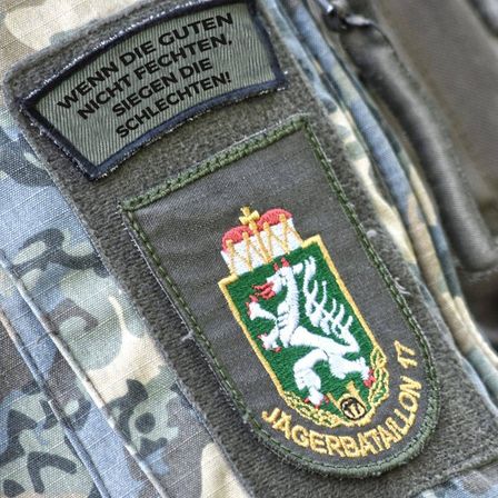 Ein Tarnmuster-Militäruniform-Aufnäher mit einem weißen Hirsch auf grünem Hintergrund, goldener Krone und den Worten 'Jaegerbataillon 1'. Darüber ein Aufnäher mit dem deutschen Text 'Wenn die Guten, nicht fechten, siegen die Schlechten!'.