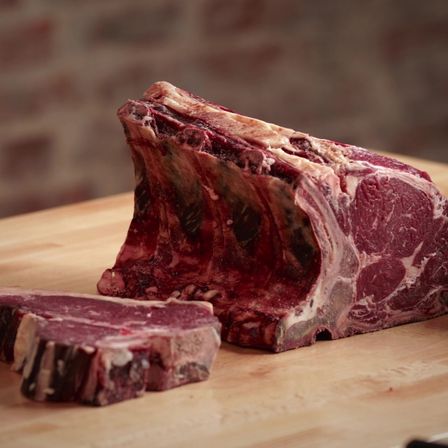 Ein roher Ribeye-Steak auf einem hölzernen Schneidebrett mit einer Ziegelwand im Hintergrund. Der Steak hat eine rotbraune Farbe.