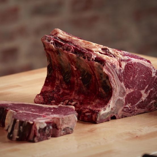 Ein roher Ribeye-Steak auf einem hölzernen Schneidebrett mit einer Ziegelwand im Hintergrund. Der Steak hat eine rotbraune Farbe.