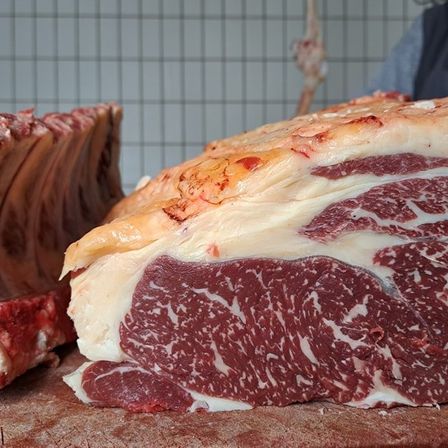 Ein großer Rinderbraten ist auf einem Holzschneidebrett zu sehen, der seine marmorierte Textur präsentiert. In der Nähe ist ein Rippenabschnitt sichtbar.