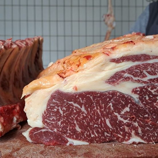 Ein großer Rinderbraten ist auf einem Holzschneidebrett zu sehen, der seine marmorierte Textur präsentiert. In der Nähe ist ein Rippenabschnitt sichtbar.