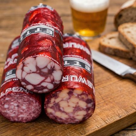 Drei rote Alpen-Wurstwaren mit einem Bier im Hintergrund auf einem Holztisch mit einem Messer und Brot.