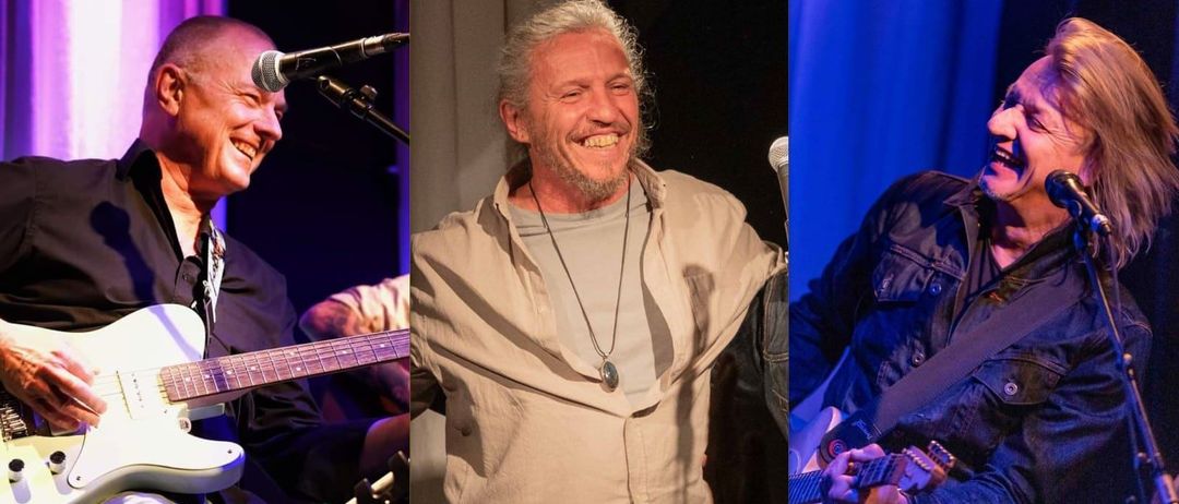 Drei Musiker sind auf der Bühne, einer spielt Gitarre, ein anderer lächelt in die Kamera und der dritte spielt Gitarre unter einem Scheinwerfer.