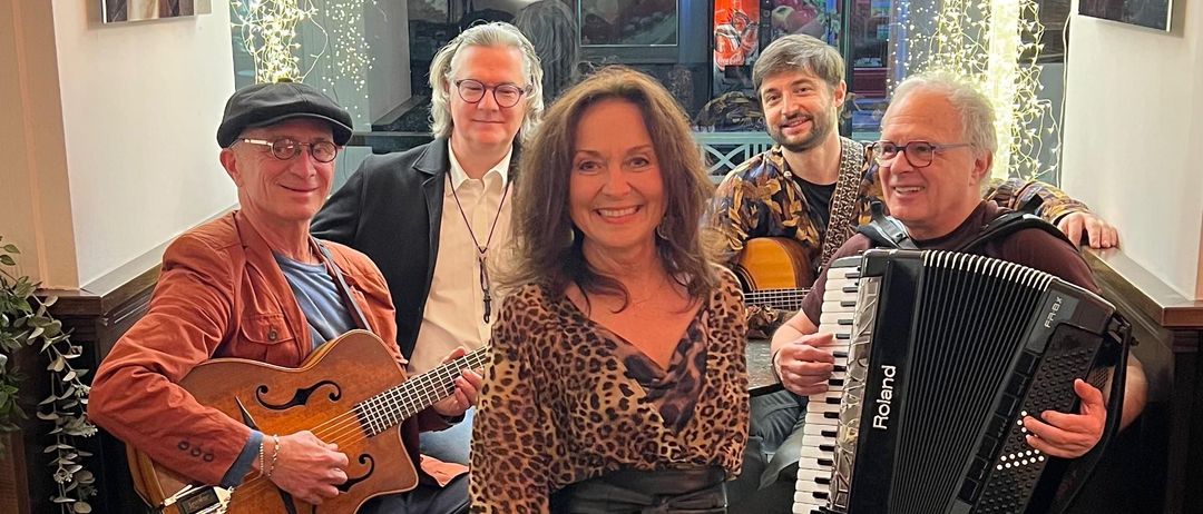 Fünf Musiker posieren für ein Foto. Eine Frau in einem Leopardenkleid steht in der Mitte. Ein Mann links spielt Gitarre. Ein anderer Mann rechts spielt Akkordeon. Sie lächeln.