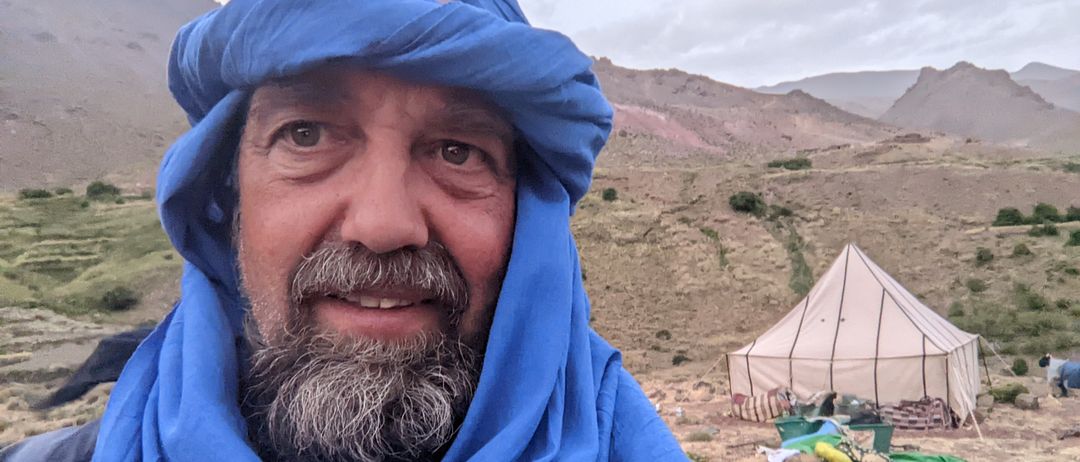 Ein Mann mit blauem Kopftuch und Bart lächelt in einer bergigen Landschaft mit einem Zelt im Hintergrund.