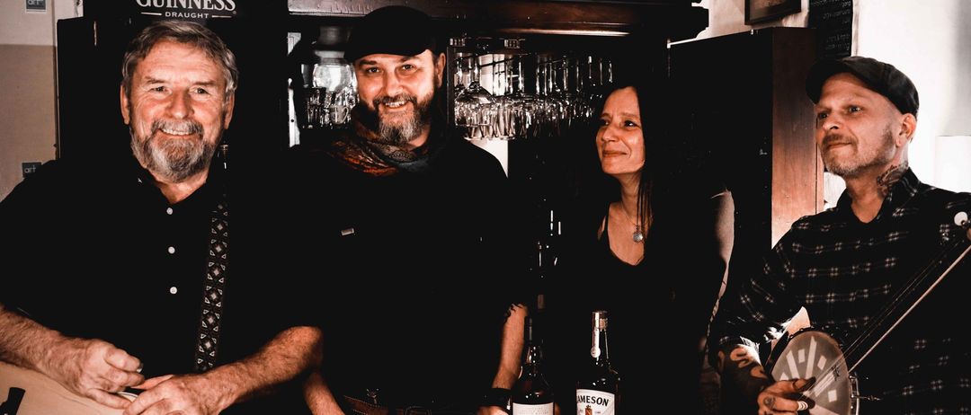 Vier Personen posieren für ein Foto in einer Bar. Ein Mann spielt Gitarre, und drei andere lächeln. Zwei Flaschen Jameson stehen auf einem Fass zwischen ihnen. Ein Guinness-Schild ist an der Wand hinter ihnen.