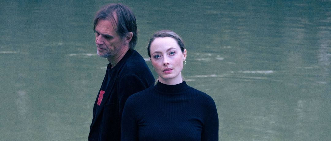 Ein Mann und eine Frau stehen auf Felsen neben einem Fluss, beide in Schwarz gekleidet. Der Mann hat einen roten Aufnäher auf seiner Jacke, während die Frau ein Rollkragenshirt und Hose trägt.