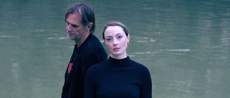 Ein Mann und eine Frau stehen auf Felsen neben einem Fluss, beide in Schwarz gekleidet. Der Mann hat einen roten Aufnäher auf seiner Jacke, während die Frau ein Rollkragenshirt und Hose trägt.