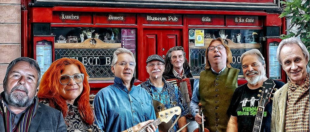 Sechs Musiker stehen vor einem roten Gebäude, möglicherweise einer Bar, namens Sean Tierney. Die Musiker halten verschiedene Instrumente, darunter ein Akkordeon, eine Geige und eine Gitarre.