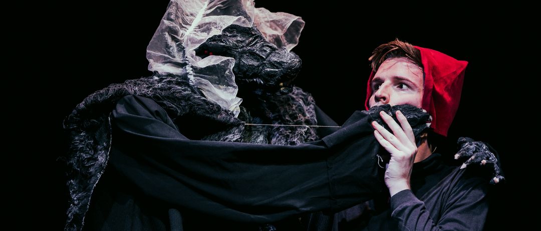 Ein Mann in einer roten Kapuze hält den Umhang eines großen, schwarzen, pelzigen Wesens und betrachtet ihn aufmerksam. Das Wesen hat eine Plastikfolie um den Kopf.