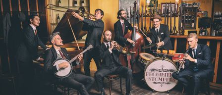 Eine Gruppe von Musikern in Anzügen spielt Instrumente. Einer spielt ein Posaune, ein anderer eine Trompete, ein dritter ein Banjo, und andere spielen Cello und Schlagzeug.