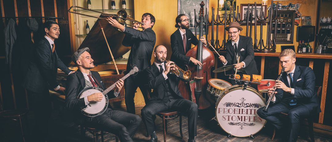 Eine Gruppe von Musikern in Anzügen spielt Instrumente. Einer spielt ein Posaune, ein anderer eine Trompete, ein dritter ein Banjo, und andere spielen Cello und Schlagzeug.