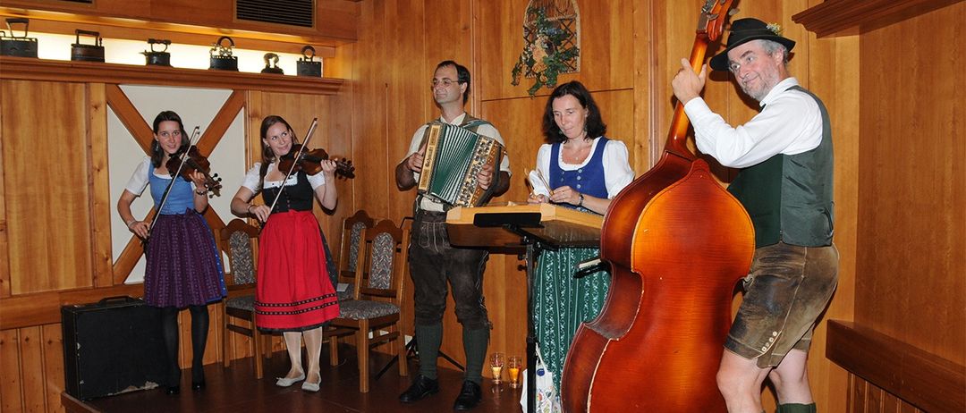 Eine Gruppe von Musikern in traditioneller bayerischer Kleidung spielt auf der Bühne. Eine Frau spielt Geige, ein Mann Akkordeon und eine andere Frau Harfe. Ein Mann spielt Cello, während ein anderer hinter ihm steht. Hinter ihnen befindet sich eine Holzwand mit Stühlen und einem Regal mit Gegenständen.